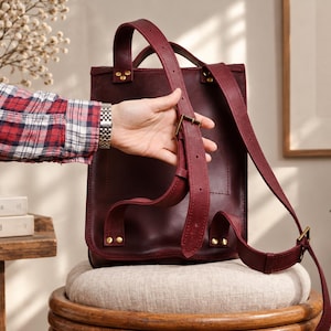 May include: A deep burgundy leather backpack featuring adjustable shoulder straps with metal buckles and reinforced rivets. The bag has a vertical rectangular structure with a top handle and a smooth, matte finish on the leather surface.