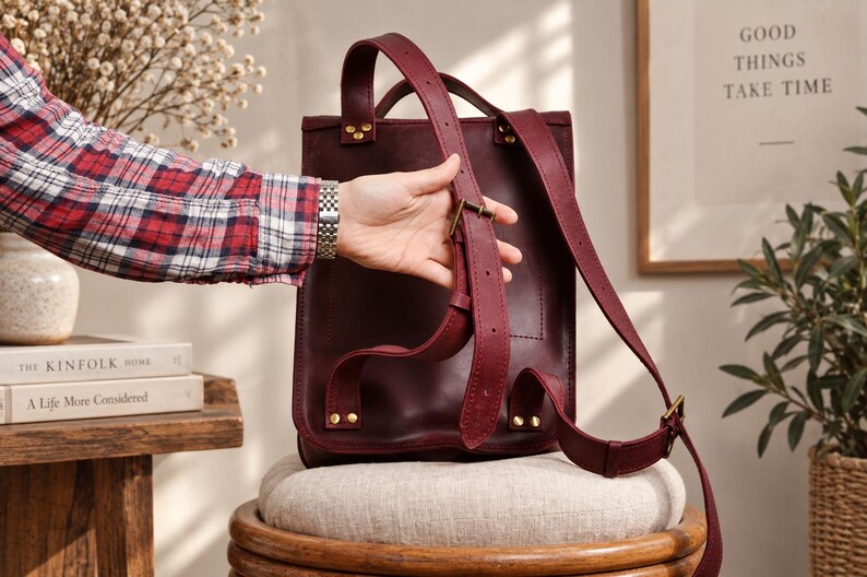 May include: A deep burgundy leather backpack featuring adjustable shoulder straps with metal buckles and reinforced rivets. The bag has a vertical rectangular structure with a top handle and a smooth, matte finish on the leather surface.