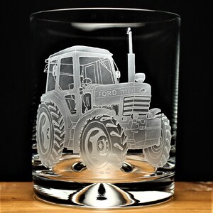 May include: A clear glass tumbler with an etched design of a Ford 7710 tractor. The tractor is facing forward with the front wheels visible. The glass has a rounded bottom.