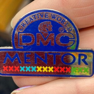 May include: A silver and blue metal pin with the text "Creative World DMC Mentor" and a row of colorful X's.