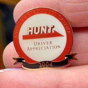 May include: A gold-colored pin with a red and white circle. The circle has the text "American Specialized Carrier" and "Hunt Transportation" in a red banner. The text "Driver Appreciation 2004" is also on the pin.
