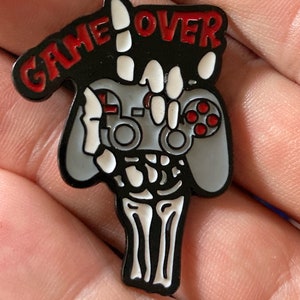 May include: A black enamel pin with a skeleton hand holding a video game controller. The hand is giving the 'peace' sign and the words 'GAME OVER' are written above the hand.