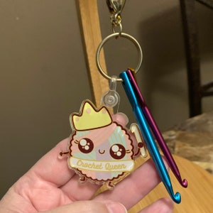 Op de afbeelding: Een transparante acryl sleutelhanger met een cartoon garenbal die een kroon draagt en een haaknaald vasthoudt. De garenbal is roze, blauw en wit met een roze en wit gestreept lint waarop "Crochet Queen" staat. De sleutelhanger is bevestigd aan een zilveren sleutelring met twee haaknaalden.