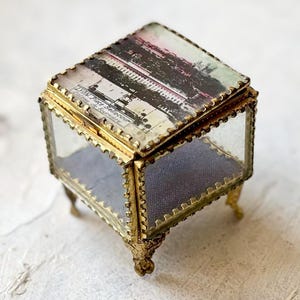 Puede incluir: Una pequeña caja de cristal de estilo vintage con un marco de metal dorado y patas ornamentadas. La tapa presenta una imagen descolorida y multicolor. El interior tiene un forro de tela azul claro, creando una pieza de almacenamiento decorativa.