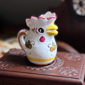 以下が含まれることがあります： 黄色いクチバシ、赤い肉垂、黒い目を持つ、鶏の顔のデザインの白いセラミックピッチャー。ピッチャーは花のモチーフで装飾されており、彫刻された花の模様の濃い茶色の木箱の上に置かれています。箱の長さは約15cmです。