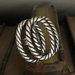 Può includere: Un anello d'argento con un design a corda. L'anello è composto da tre bande intrecciate, ciascuna con una texture a corda in rilievo.