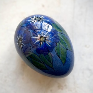 May include: A blue, egg-shaped decorative object with a glossy finish. The surface is adorned with a floral design featuring blue flowers and green leaves. The egg is a solid color with a smooth texture.