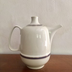 Op de afbeelding: Een witte theepot van keramiek met een gouden rand en een blauwe en witte band met een patroon rond het midden. De theepot heeft een rond lichaam en een tuit aan de zijkant.