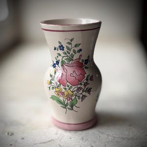May include: A cream ceramic vase with a pink floral design. The vase has a pink base and rim, with a large pink rose and smaller blue and yellow flowers. The design is hand-painted and the vase has a classic shape.