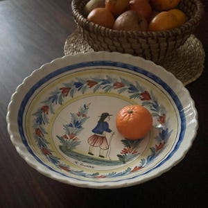 Può includere: Una ciotola da portata in ceramica bianca con bordo smerlato, con un disegno dipinto a mano di una persona in abiti tradizionali e motivi floreali. Un'arancia è all'interno della ciotola. Un cestino di vimini con frutta è sullo sfondo.
