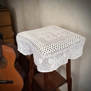 May include: A white, square, crocheted table cover with a geometric pattern, draped over a small, dark wooden table. The cover has a scalloped edge. A classical guitar is visible in the background.