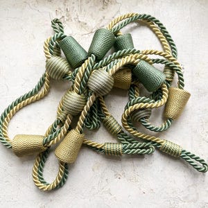 Può includere: Una collezione di fermatende decorative nei toni del verde e dell'oro. I fermatende presentano cordoncini di corda ritorta e accenti cilindrici strutturati. Il design include varie tonalità di verde e oro, creando un look sofisticato.
