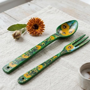 Può includere: Un set di posate da insalata in legno composto da un cucchiaio e una forchetta, entrambi dipinti in verde scuro e decorati con motivi di girasoli gialli e fogliame verde dipinti a mano. Ogni manico presenta un piccolo foro circolare.