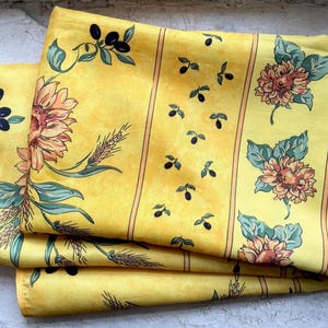 Könnte beinhalten: Gefalteter gelber Stoff mit einem Blumen- und Olivenzweigmuster. Das Design zeigt Sonnenblumen, Weizen und Olivenzweige in Orange-, Grün- und Schwarztönen. Der Stoff hat vertikale Streifen.