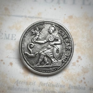 Puede incluir: Una moneda de color plateado que representa una figura sentada con vestimenta clásica, sosteniendo un pájaro y un escudo. La moneda está inscrita con la palabra "CAESAR". La moneda tiene un diseño antiguo detallado.