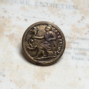 Puede incluir: Botón redondo de bronce antiguo con un relieve detallado de una figura sentada sosteniendo un pájaro y una serpiente. El botón tiene un borde en relieve y texto alrededor del borde, lo que sugiere un origen histórico.