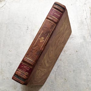 Op de afbeelding: Antiek hardcover boek met een bruine leren rug en een gemarmerde kaft. De rug heeft gouden letters en decoratieve reliëfdetails. De titel "V. HUGO" is zichtbaar, samen met andere tekst. De randen van het boek zijn versleten.