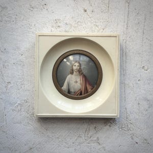 Puede incluir: Imagen religiosa enmarcada que presenta un retrato de Jesucristo. La imagen está dentro de un marco circular con un borde de color bronce, encerrada en un marco cuadrado de plástico blanquecino. El retrato muestra a Jesús con pelo largo, túnica blanca y chal rojo.