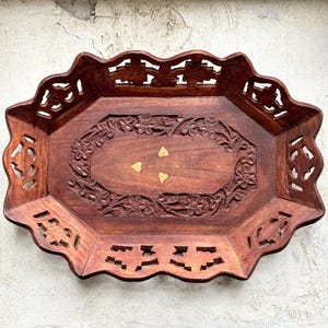 May include: A dark brown wooden decorative tray with detailed floral carvings and cut-out patterns. The oval tray has scalloped edges and small gold heart-shaped accents. The tray is likely used for serving or display, and is made of wood.