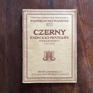 May include: A vintage book cover with a brown border and text in French. The title is "CZERNY" and the subtitle is "EXERCICES PRATIQUES POUR LES DOIGTS". The book is part of the "PANTHEON-DES-PIANISTES" series.