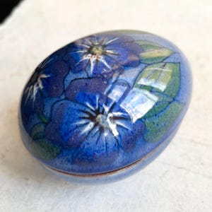 Puede incluir: Caja de baratijas ovalada de cerámica con acabado brillante. La caja es principalmente azul, adornada con un estampado floral con flores azules, hojas verdes y detalles blancos. La tapa y la base son separadas.
