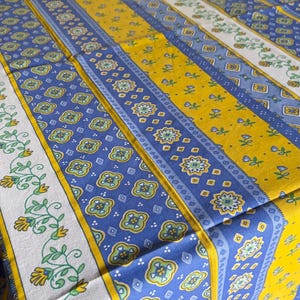 May include: A rectangular tablecloth featuring a repeating pattern of vertical stripes in vibrant yellow, deep blue, and white. The design includes intricate floral motifs, geometric shapes, and scrolling vine patterns printed across the fabric surface.