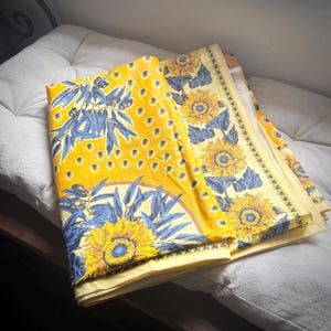 Op de afbeelding: Gele stof met een zonnebloem- en olijftakontwerp. De stof heeft een herhalend patroon van zonnebloemen en blauwe bladeren op een gele achtergrond. De stof is gevouwen en ligt op een wit oppervlak.