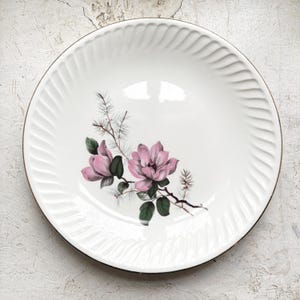 Könnte beinhalten: Weißer Keramikteller mit Goldrand und geriffeltem Rand. Der Teller zeigt ein Blumendesign aus rosa Magnolienblüten und grünen Blättern. Der Teller ist ein dekoratives Stück.