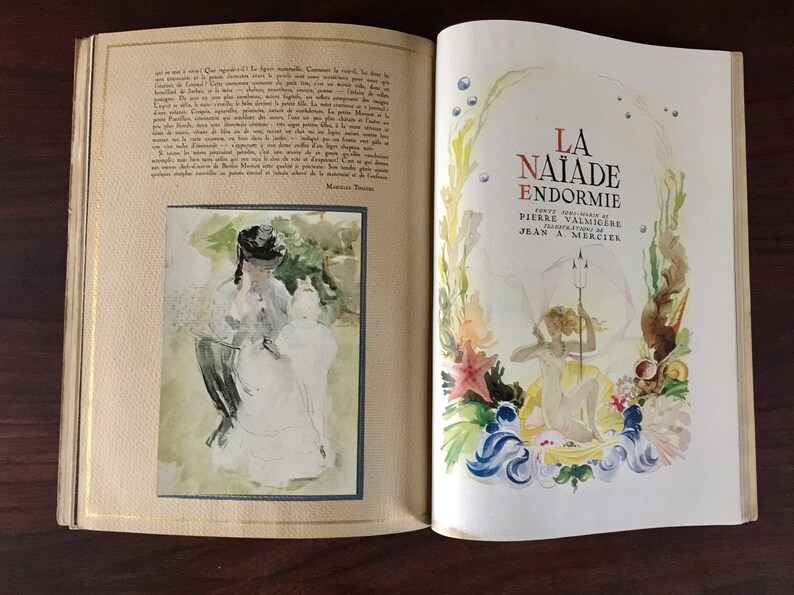 May include: An open book with a watercolor illustration of a woman and a child on the left page. The right page features a watercolor illustration of a mermaid with a trident, surrounded by seaweed and coral. The title of the book is "La Naiade Endormie" with the author's name, Pierre Valmig&egrave;re, and the illustrator's name, Jean A. Mercier.