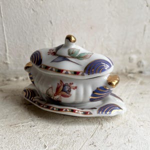 Può includere: Un portagioie in porcellana bianca con coperchio, con motivi floreali e geometrici blu, rossi, verdi e dorati. La scatola poggia su un piattino abbinato. Il coperchio ha un pomello dorato.