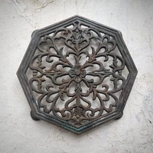 Op de afbeelding: Oud ijzeren onderzetter met een donkere, verweerde afwerking. De zeshoekige onderzetter heeft een centraal bloemmotief, omringd door sierlijke, opengewerkte patronen. Voorzien van kleine voetjes. Ongeveer 20 cm in diameter.