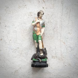 May include: A detailed figurine of St. Michael, in armour and holding a spear, standing over a defeated dark figure. The statue features green, orange, and silver accents, with the base inscribed "ST MICHEL".