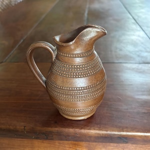 May include: A brown ceramic pitcher with a handle and a spout. The pitcher has a series of horizontal bands with a dotted pattern. The pitcher is sitting on a wooden surface.