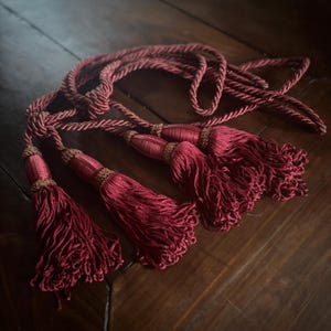 Puede incluir: Borlas decorativas de color burdeos con un diseño de cuerda trenzada. Las borlas tienen una apariencia completa y fluida y están acentuadas con un detalle marrón más oscuro en la parte superior. La cuerda también es burdeos, creando un aspecto cohesivo. Las borlas están dispuestas sobre una superficie de madera oscura.