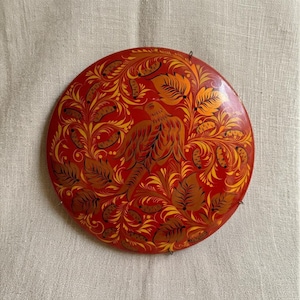May include: A circular red decorative plate featuring a hand-painted golden and brown bird surrounded by intricate swirling floral patterns, leaves, and small seed-like shapes. The surface has a glossy finish with a metal wire hanger.