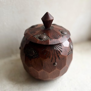Puede incluir: Un recipiente de madera marrón oscuro con tapa y un diseño geométrico facetado. La tapa presenta motivos tallados de piñas y hojas. El recipiente tiene un pomo en forma de diamante y pequeños pernos metálicos. La forma general es redonda.