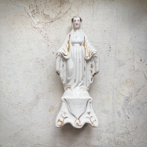 May include: A white porcelain statue of a religious figure with gold accents. The figure is standing with arms outstretched, wearing a flowing robe. The statue is mounted on a decorative base.