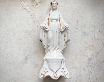 Puede incluir: Estatua de porcelana blanca de una figura religiosa con detalles dorados. La figura está de pie con los brazos extendidos, vestida con una túnica. La estatua está montada sobre una base decorativa.