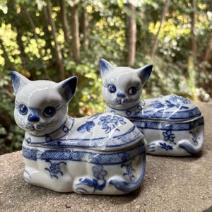 以下が含まれることがあります： 青い手描きの花柄と波模様が施された、白い猫の形をした陶器製の小物入れ2つ。それぞれの猫は、尖った耳、青い目、首輪のディテールを持ち、横たわった姿勢で描かれています。上半分は取り外し可能な蓋になっています。