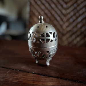Può includere: Un bruciatore di incenso argentato, di forma sferica, con motivi decorativi traforati e un piccolo pomello sulla parte superiore. Il bruciatore ha tre piedini e poggia su una superficie di legno scuro. Lo sfondo è sfocato.