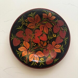Könnte beinhalten: Ein runder, schwarzer Dekoteller mit einem handgemalten Motiv aus roten Erdbeeren sowie orangefarbenen und grünen Blättern. Das Design enthält kleine gelbe florale Akzente, die in der Mitte der dunklen Oberfläche angeordnet sind.