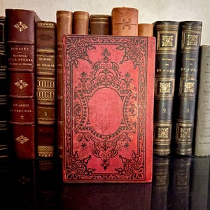 May include: A selection of antique books with leather covers. The central book is red with a black decorative pattern. Other books are in shades of brown and black, some with gold detailing. The books are displayed on a dark surface.