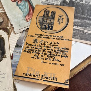 May include: A rectangular tan paper card featuring a black ink illustration of Notre-Dame de Paris, a monogram, and French text. The card includes the date Paris - 13 juillet 1937 and the name cardinal Pacelli printed in a decorative black font.