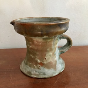May include: A brown and green ceramic pitcher with a handle. The pitcher has a wide mouth and a narrow base.