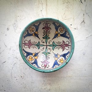 Puede incluir: Un cuenco redondo de cerámica pintado a mano, con interior blanco y borde turquesa. El cuenco presenta un diseño simétrico con pájaros azules, flores amarillas y elementos florales morados y verdes. Probablemente se utiliza para servir alimentos o como pieza decorativa.