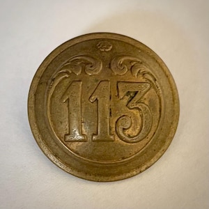 Puede incluir: Un botón redondo de color latón antiguo con el número "113" en el centro. Los números están en relieve y rodeados de adornos decorativos. Un pequeño diseño floral en la parte superior.