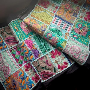 May include: A detailed patchwork textile with vibrant embroidery. The design incorporates squares of teal, pink, and gold, featuring floral and paisley patterns. A light blue border trims the edges, creating a colourful and intricate piece, suitable for decorative use.