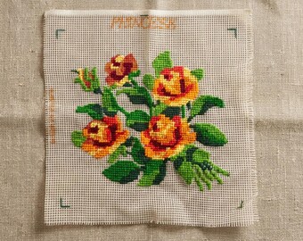 May include: A square piece of beige Aida fabric featuring a cross-stitch pattern of orange and yellow roses with green leaves. The word PRINCESSA is stitched in orange at the top centre, and green corner registration marks are visible.
