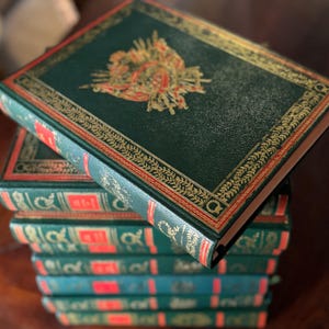 Op de afbeelding: Een stapel antieke boeken met donkergroene kaften. De kaften hebben gouden en rode decoratieve randen en een gouden embleem. De ruggen zijn rood met gouden letters. De boeken zijn op een houten oppervlak gestapeld.