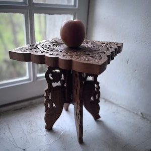 Op de afbeelding: Een kleine, ingewikkeld gesneden houten tafel met een stervormig ontwerp op het bovenste oppervlak. De tafel heeft vier poten en is gebeitst in een warme bruine kleur. Een rode appel rust op het tafeloppervlak. De tafel staat bij een raam.